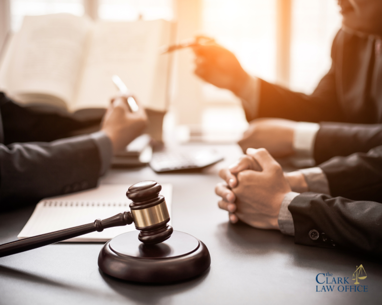 A gavel, notebook, and three people in business attire sit at a table discussing legal documents; “The Clark Law Office” logo is visible in the lower right corner. A gavel, notebook, and three people in business attire sit at a table discussing legal documents; "The Clark Law Office" logo is visible in the lower right corner.