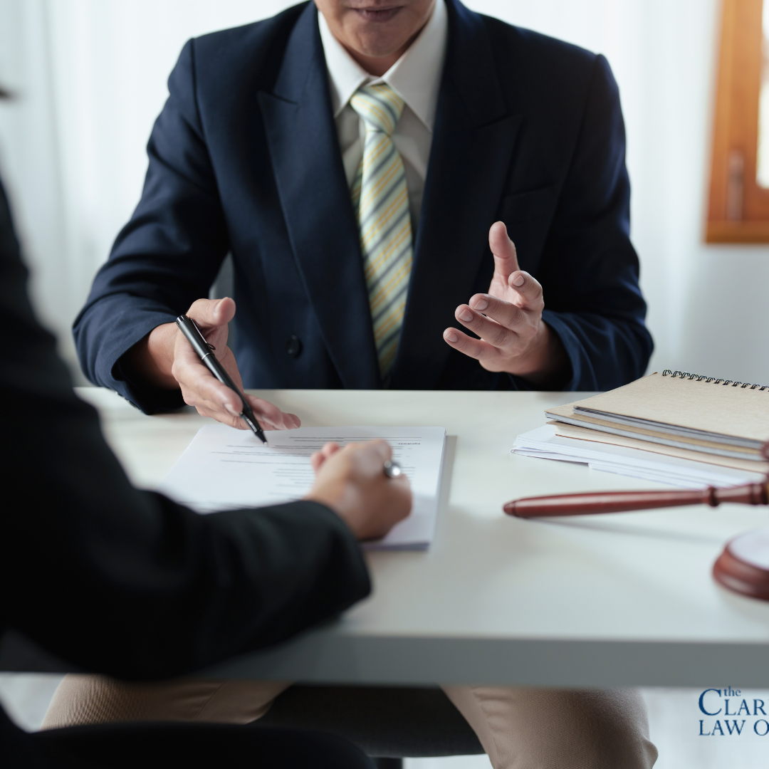 Two people sit at a desk discussing documents; one gestures while the other reviews a form. A gavel and notepad are on the table. The Clark Law Office logo is visible. Two people sit at a desk discussing documents; one gestures while the other reviews a form. A gavel and notepad are on the table. The Clark Law Office logo is visible.