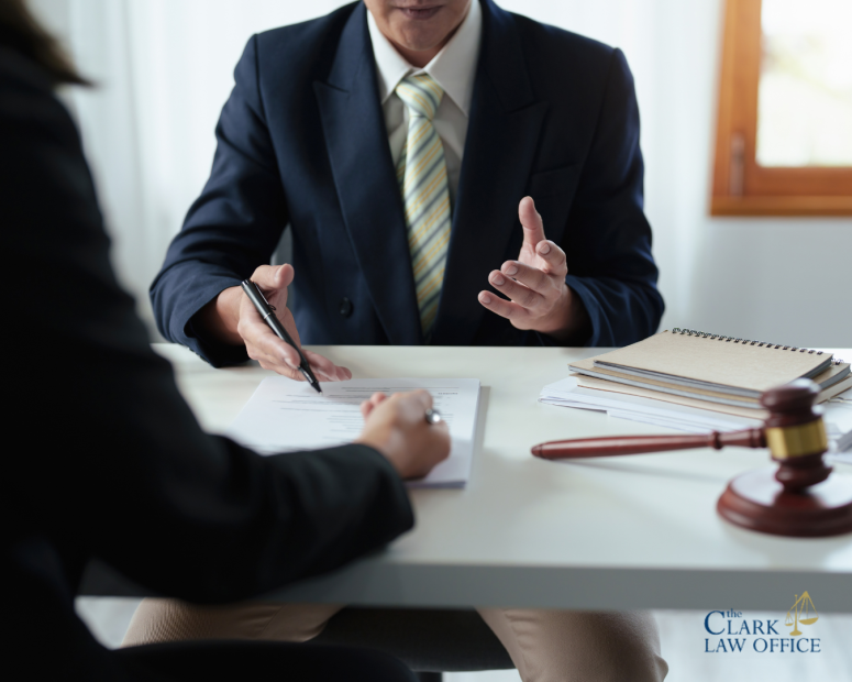 Two people sit at a desk discussing documents; one gestures while the other reviews a form. A gavel and notepad are on the table. The Clark Law Office logo is visible. Two people sit at a desk discussing documents; one gestures while the other reviews a form. A gavel and notepad are on the table. The Clark Law Office logo is visible.