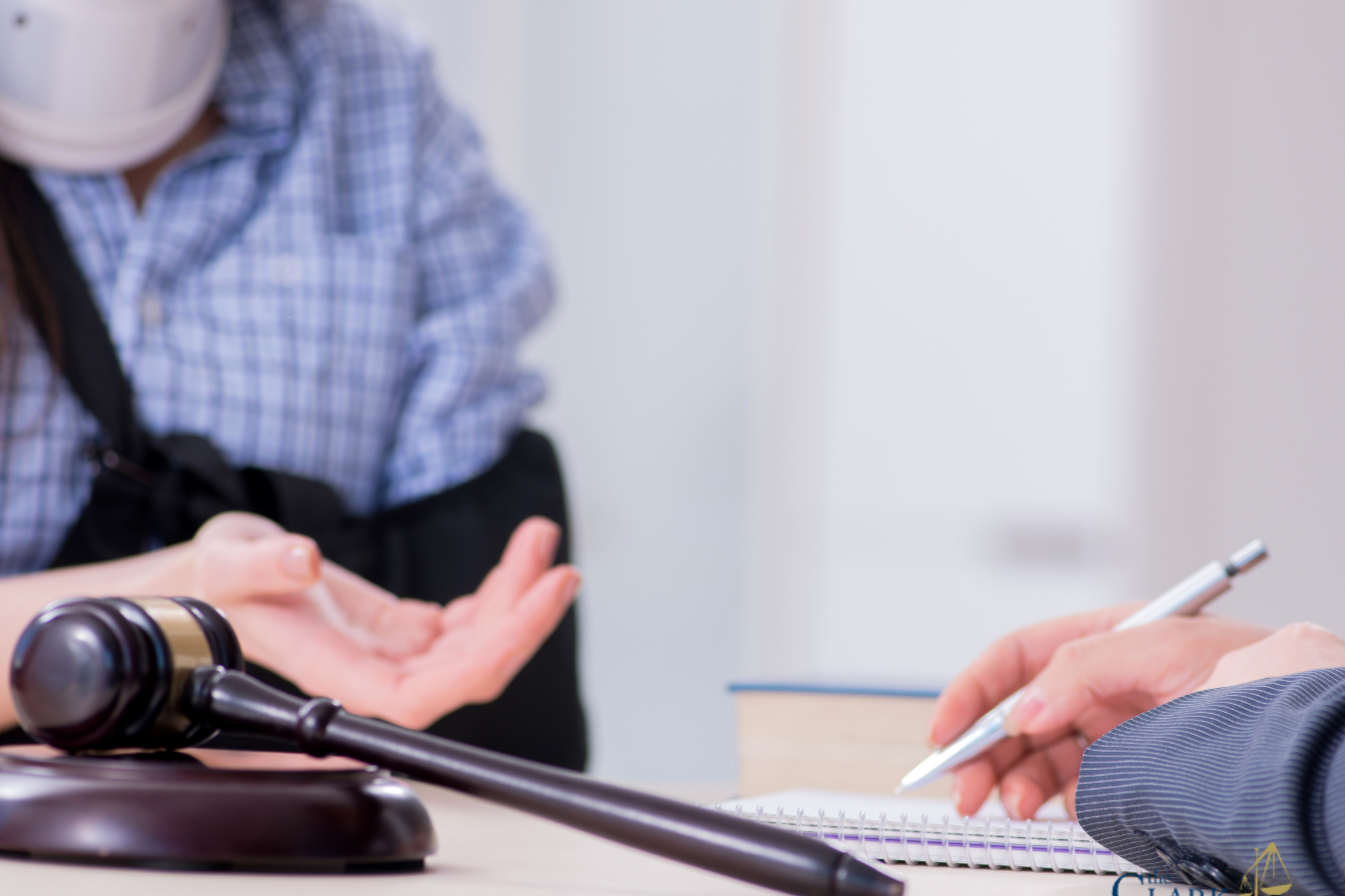 Person wearing a neck brace and arm sling sits across from a professional who is writing in a notebook; a gavel is visible on the table. Clark Law Office logo in corner.