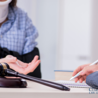 Person wearing a neck brace and arm sling sits across from a professional who is writing in a notebook; a gavel is visible on the table. Clark Law Office logo in corner.