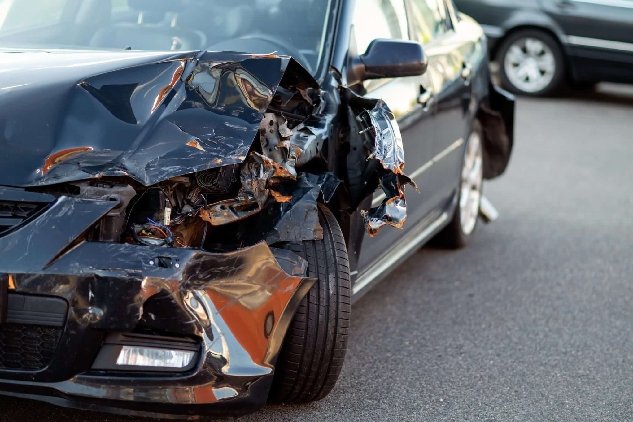 A black car with severe damage to its front left side is parked on a paved surface, suggesting it was involved in an accident. Another vehicle is visible in the background.