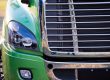 Close-up of the front end of a green semi-truck, showing the left headlight and chrome grille in detail.