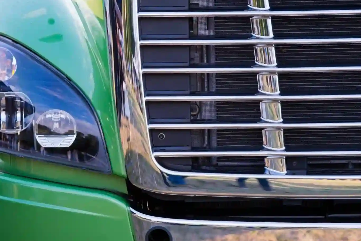 Close-up of the front left side of a green semi-truck, showing the headlight, grille, and part of the bumper. Close-up of the front left side of a green semi-truck, showing the headlight, grille, and part of the bumper.
