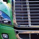 Close-up of the front left side of a green semi-truck, showing the headlight, grille, and part of the bumper.