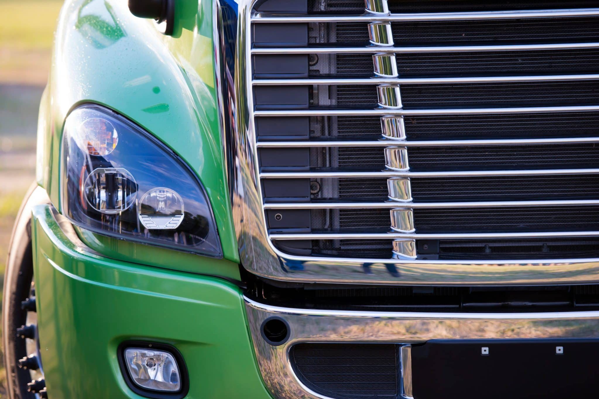 Close-up of the front end of a green semi-truck, showing the left headlight and chrome grille in detail.