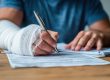 Person with a bandaged arm signing paperwork at a table, using their uninjured hand to fill out forms.
