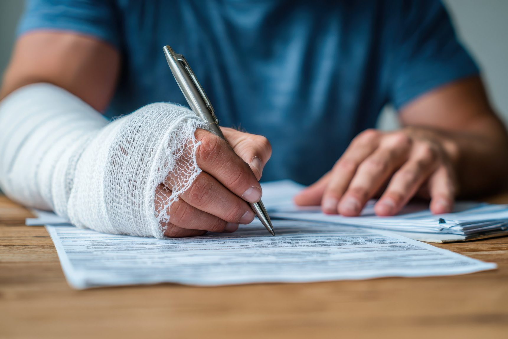 Why Do Similar Injury Cases Settle For Different Amounts Person with a bandaged arm signing paperwork at a table, using their uninjured hand to fill out forms.