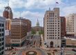 A view down a city street leading to a domed capitol building, surrounded by tall office buildings and a roundabout in the foreground.