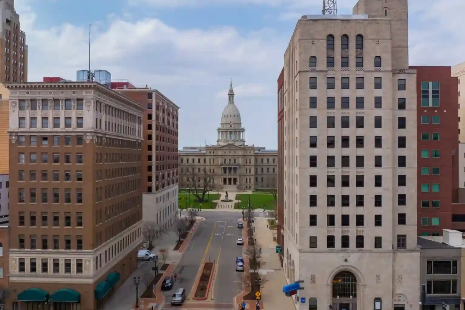 A street view in downtown Lansing, Michigan, with the Michigan State Capitol building in the background and an American flag atop a tall building.