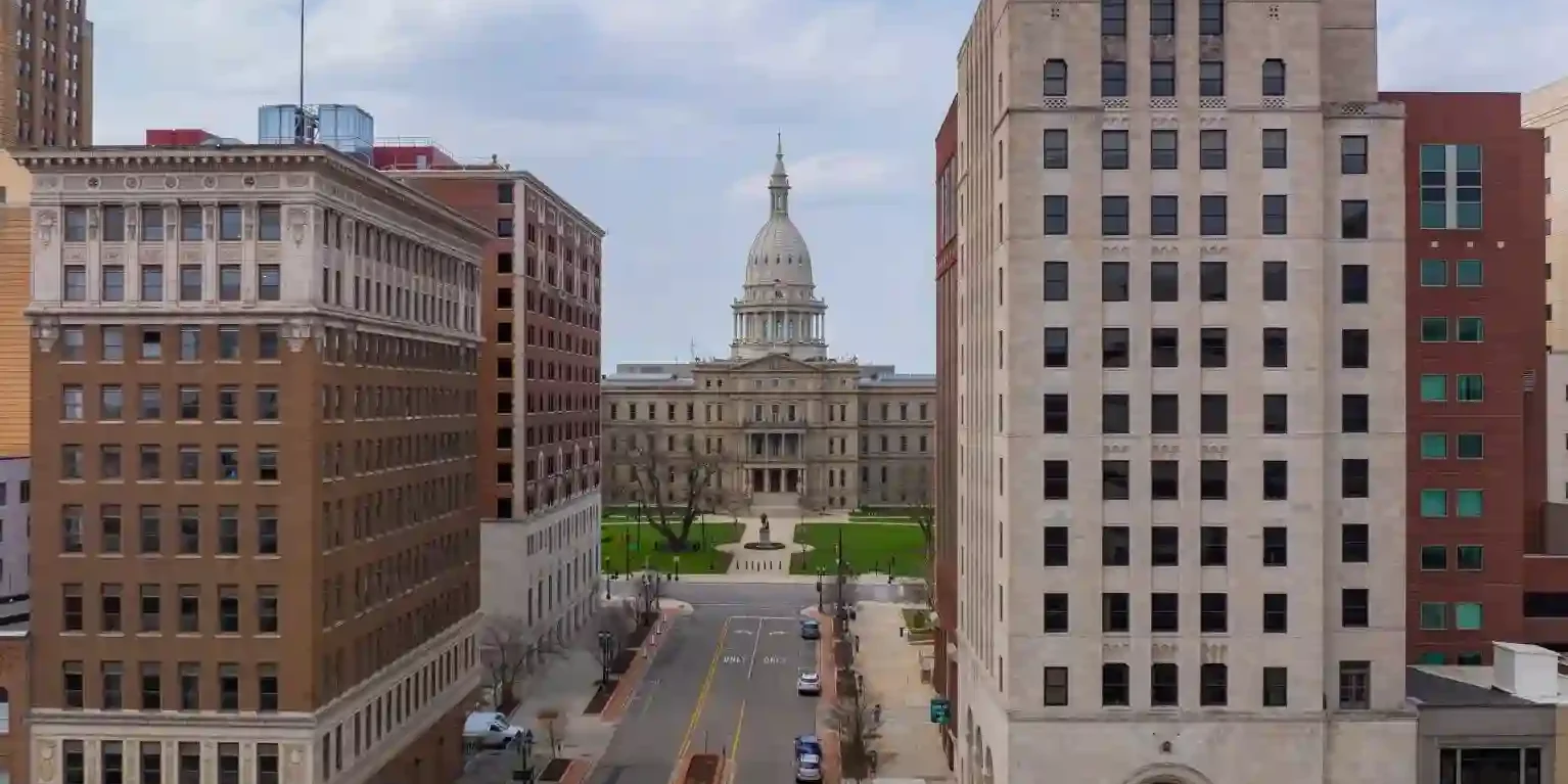 A street view in downtown Lansing, Michigan, with the Michigan State Capitol building in the background and an American flag atop a tall building. A street view in downtown Lansing, Michigan, with the Michigan State Capitol building in the background and an American flag atop a tall building.