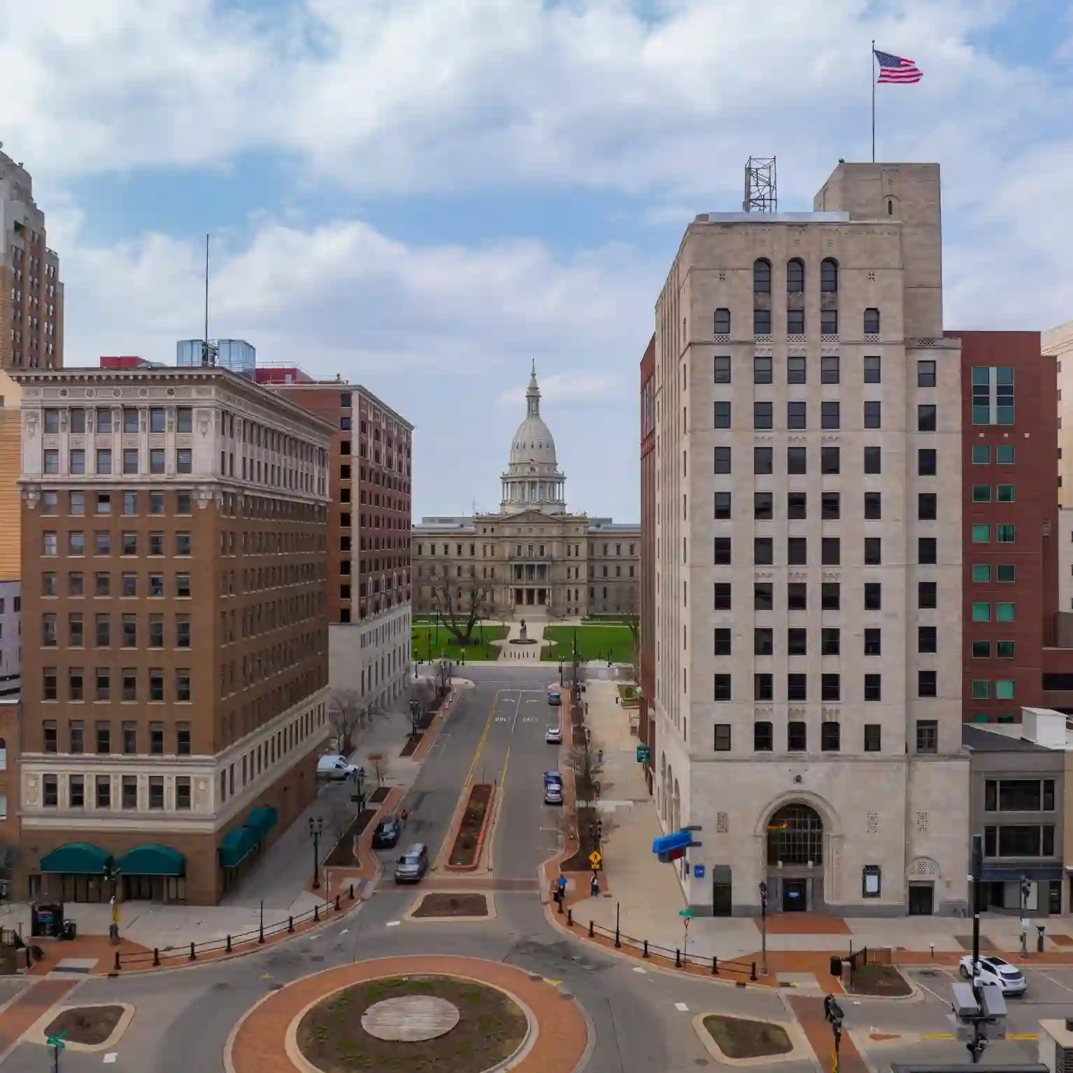 A street view in downtown Lansing, Michigan, with the Michigan State Capitol building in the background and an American flag atop a tall building.