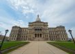 A wide-angle view of the Michigan State Capitol building with a central dome, symmetrical wings, and cloudy sky above.