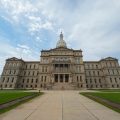 A wide-angle view of the Michigan State Capitol building with a central dome, symmetrical wings, and cloudy sky above.
