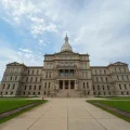 Large stone government building with a central dome and columns, viewed from a path leading up to the entrance, with a partly cloudy sky above.