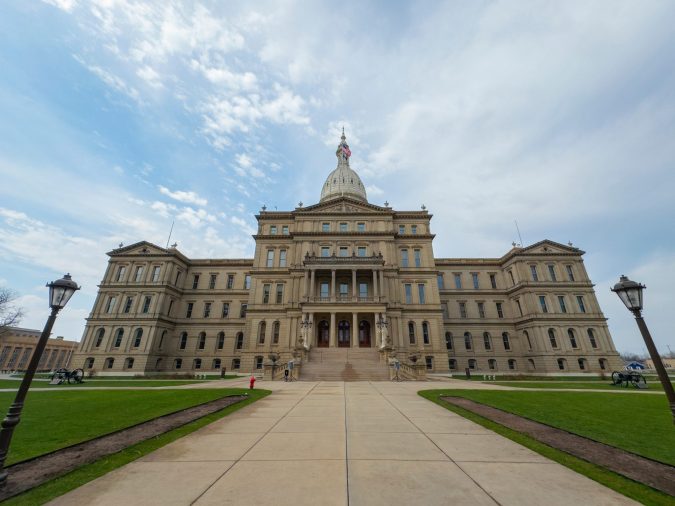 A wide-angle view of the Michigan State Capitol building with a central dome, symmetrical wings, and cloudy sky above.