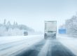 A truck drives on a snow-covered highway with trees lining the road and another vehicle approaching from the opposite direction in wintry conditions.