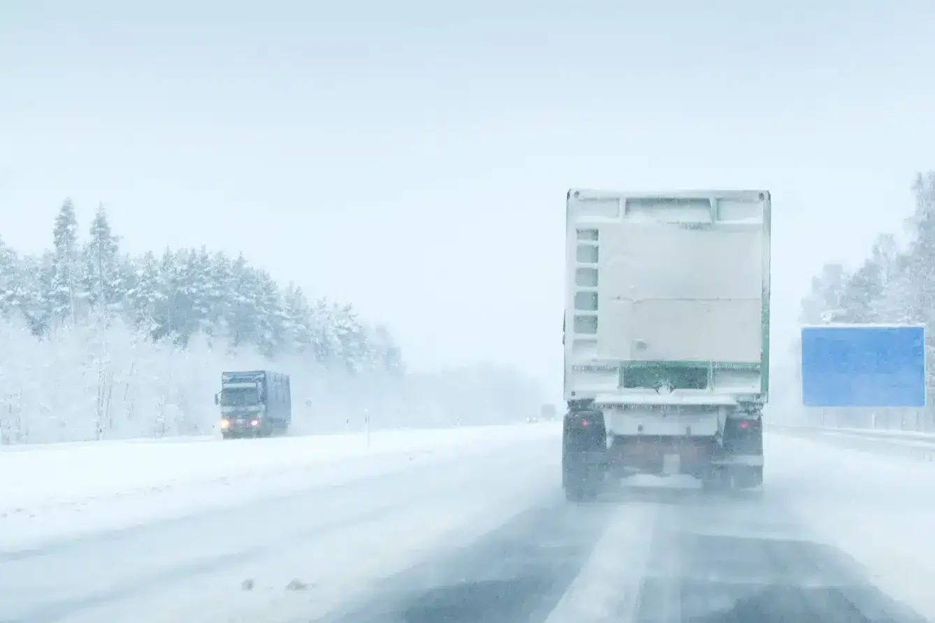 Two trucks drive on a snow-covered highway surrounded by trees, with blowing snow reducing visibility and an empty blue road sign visible on the right. Two trucks drive on a snow-covered highway surrounded by trees, with blowing snow reducing visibility and an empty blue road sign visible on the right.