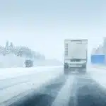 Two trucks drive on a snow-covered highway surrounded by trees, with blowing snow reducing visibility and an empty blue road sign visible on the right.