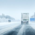 A truck drives on a snow-covered highway with trees lining the road and another vehicle approaching from the opposite direction in wintry conditions.