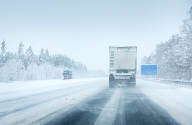 A truck drives on a snow-covered highway with trees lining the road and another vehicle approaching from the opposite direction in wintry conditions.