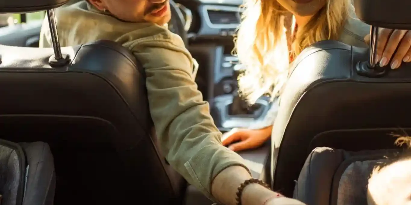 A man and woman in the front seats of a car turn around to interact with a baby seated in a rear-facing car seat in the back. A man and woman in the front seats of a car turn around to interact with a baby seated in a rear-facing car seat in the back.