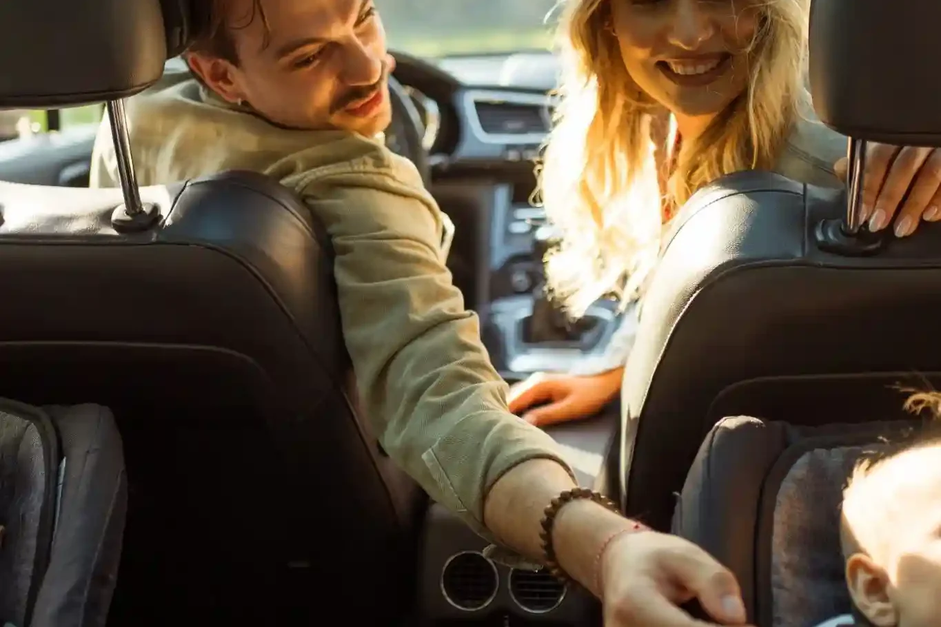 A man and woman in the front seats of a car turn around to interact with a baby seated in a rear-facing car seat in the back.