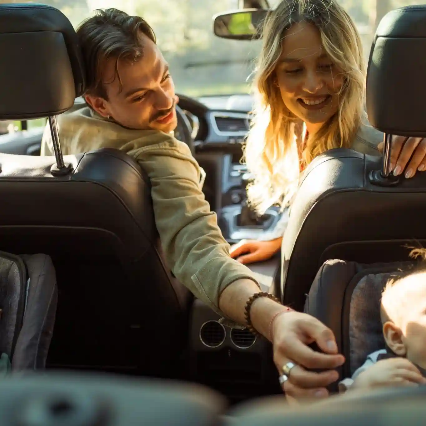 A man and woman in the front seats of a car turn around to interact with a baby seated in a rear-facing car seat in the back.