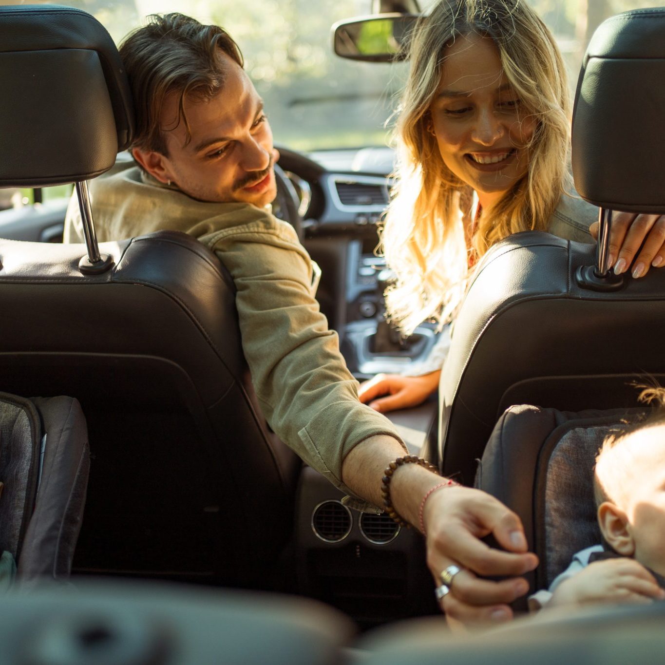 A man and woman sit in the front seats of a car, smiling and reaching toward two babies seated in car seats in the back.