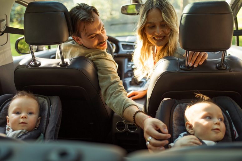 A man and woman sit in the front seats of a car, smiling and reaching toward two babies seated in car seats in the back.