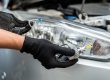 Person wearing black gloves installing or replacing a car headlight bulb, with the vehicle's headlight assembly visible in the background.
