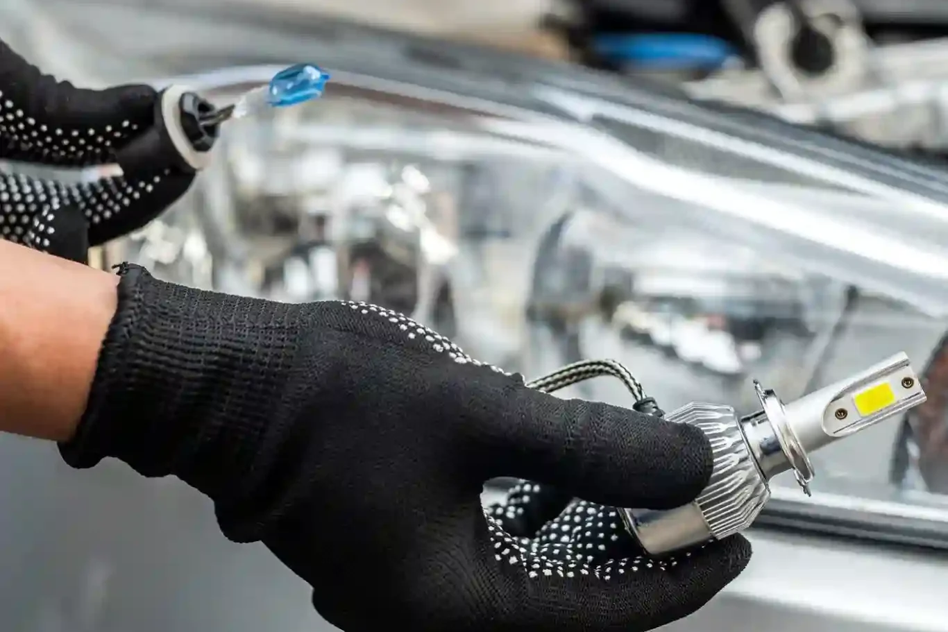 A person wearing black gloves holds an LED car headlight bulb in front of a vehicle’s headlight assembly. A person wearing black gloves holds an LED car headlight bulb in front of a vehicle's headlight assembly.
