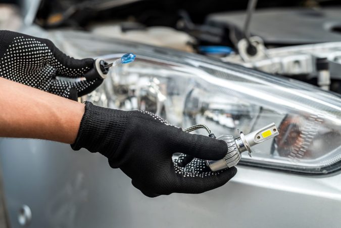 Person wearing black gloves installing or replacing a car headlight bulb, with the vehicle's headlight assembly visible in the background.