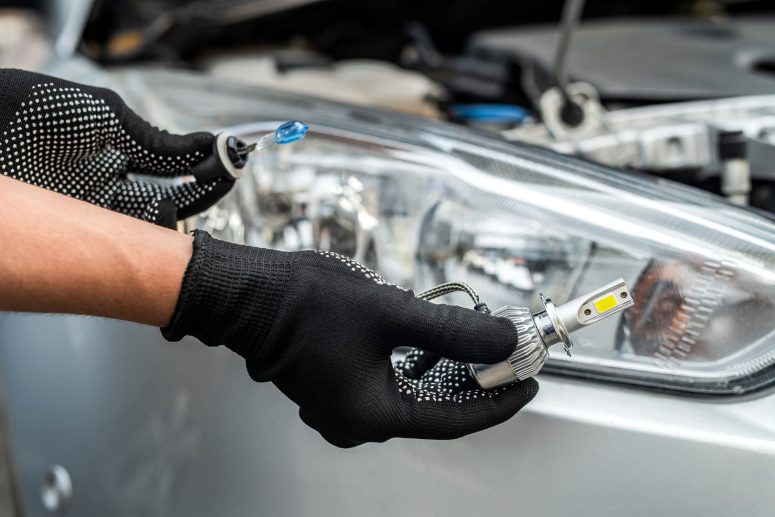 Person wearing black gloves installing or replacing a car headlight bulb, with the vehicle's headlight assembly visible in the background.