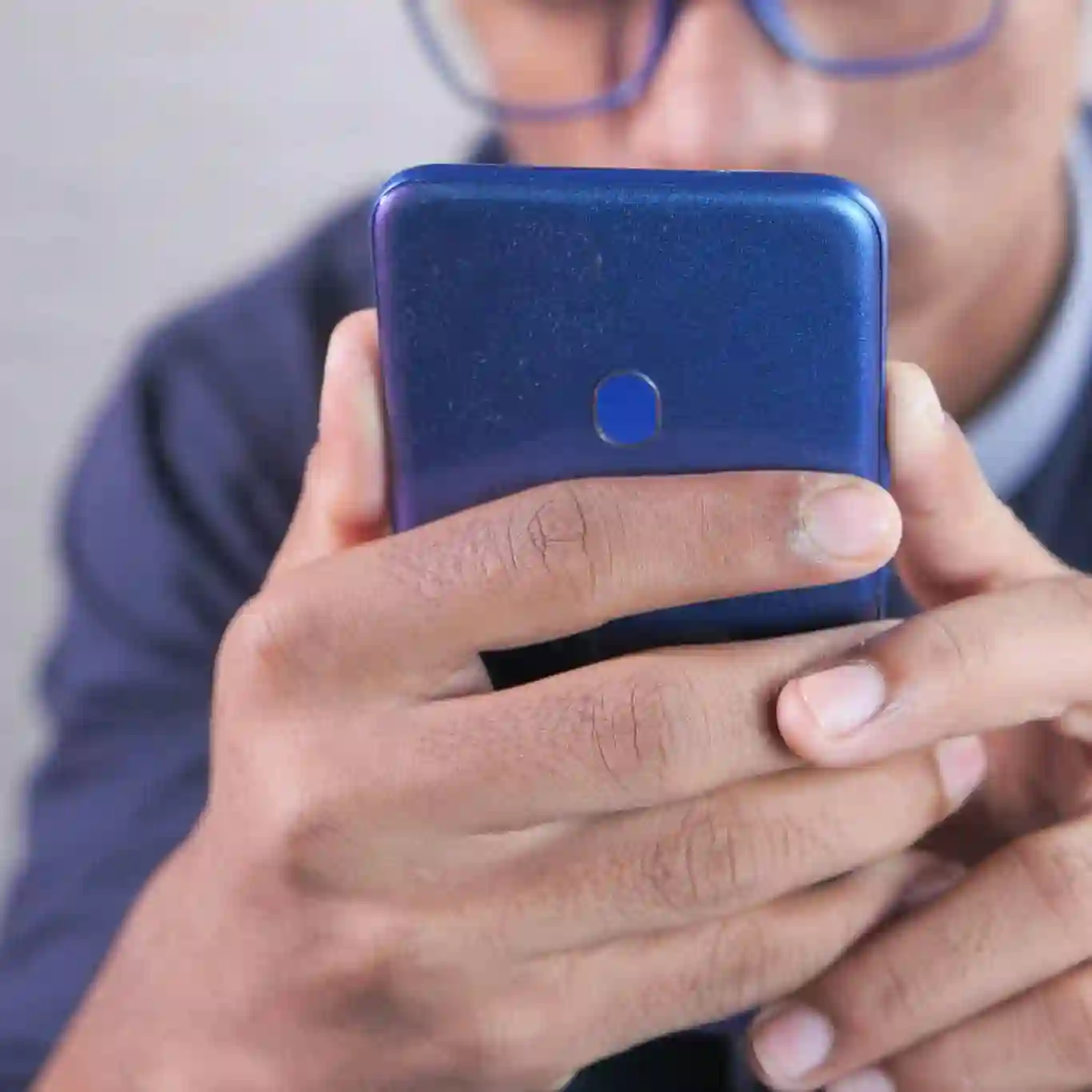 A person wearing glasses holds and looks at a blue smartphone, with their face partially visible in the background.