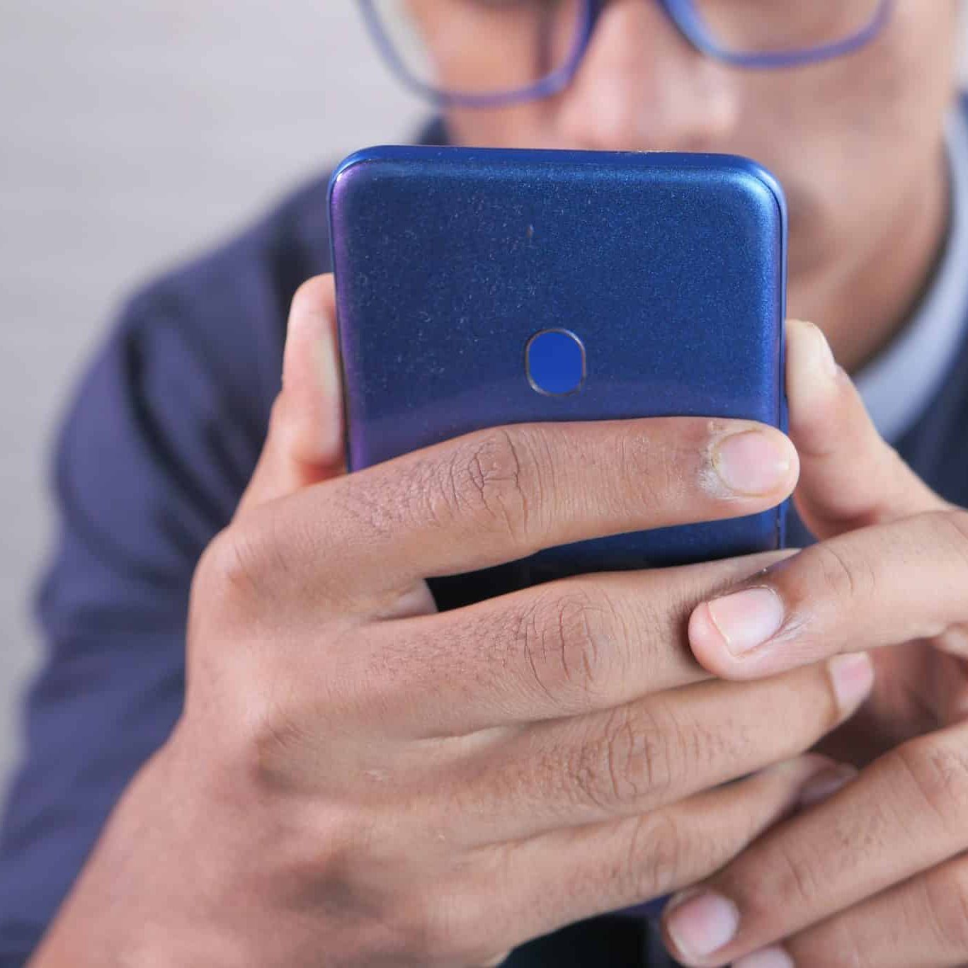 A person wearing glasses holds a blue smartphone with both hands, focusing intently on the screen.