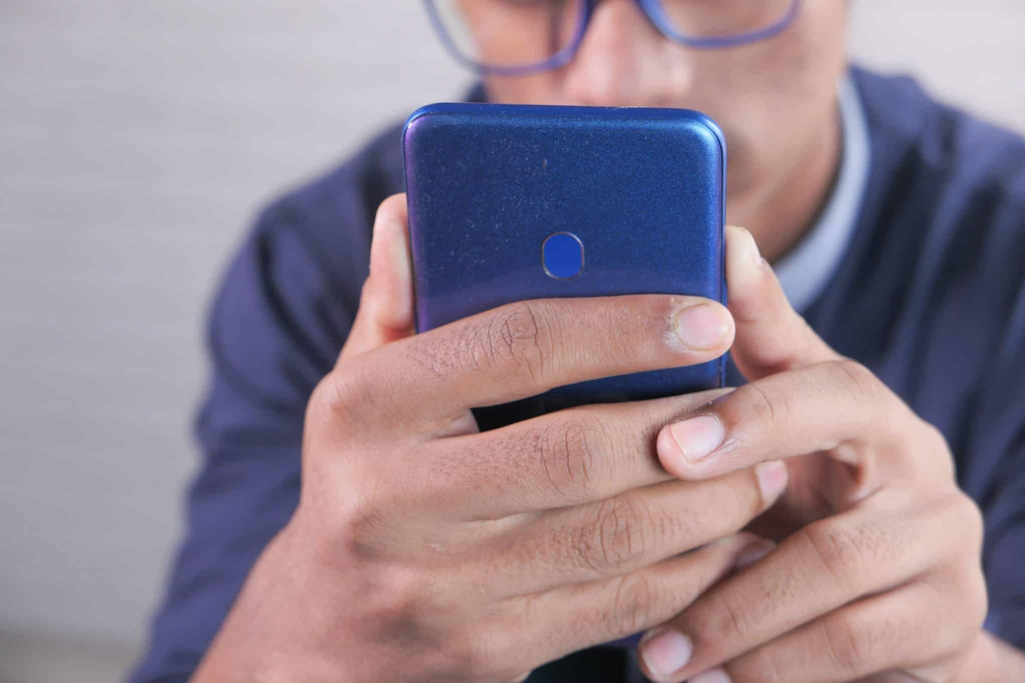 A person wearing glasses holds a blue smartphone with both hands, focusing intently on the screen.