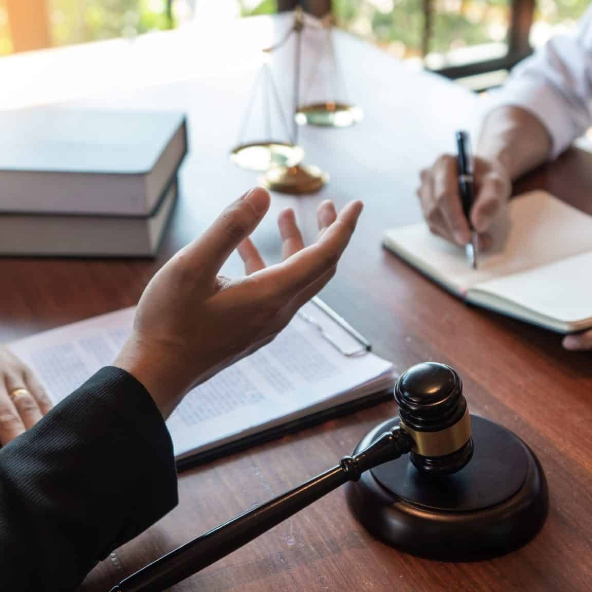 What Personal Injury Lawyers Actually Do After You Hire Them Two people sit at a desk with legal documents, a gavel, and scales of justice; one person gestures while the other takes notes.