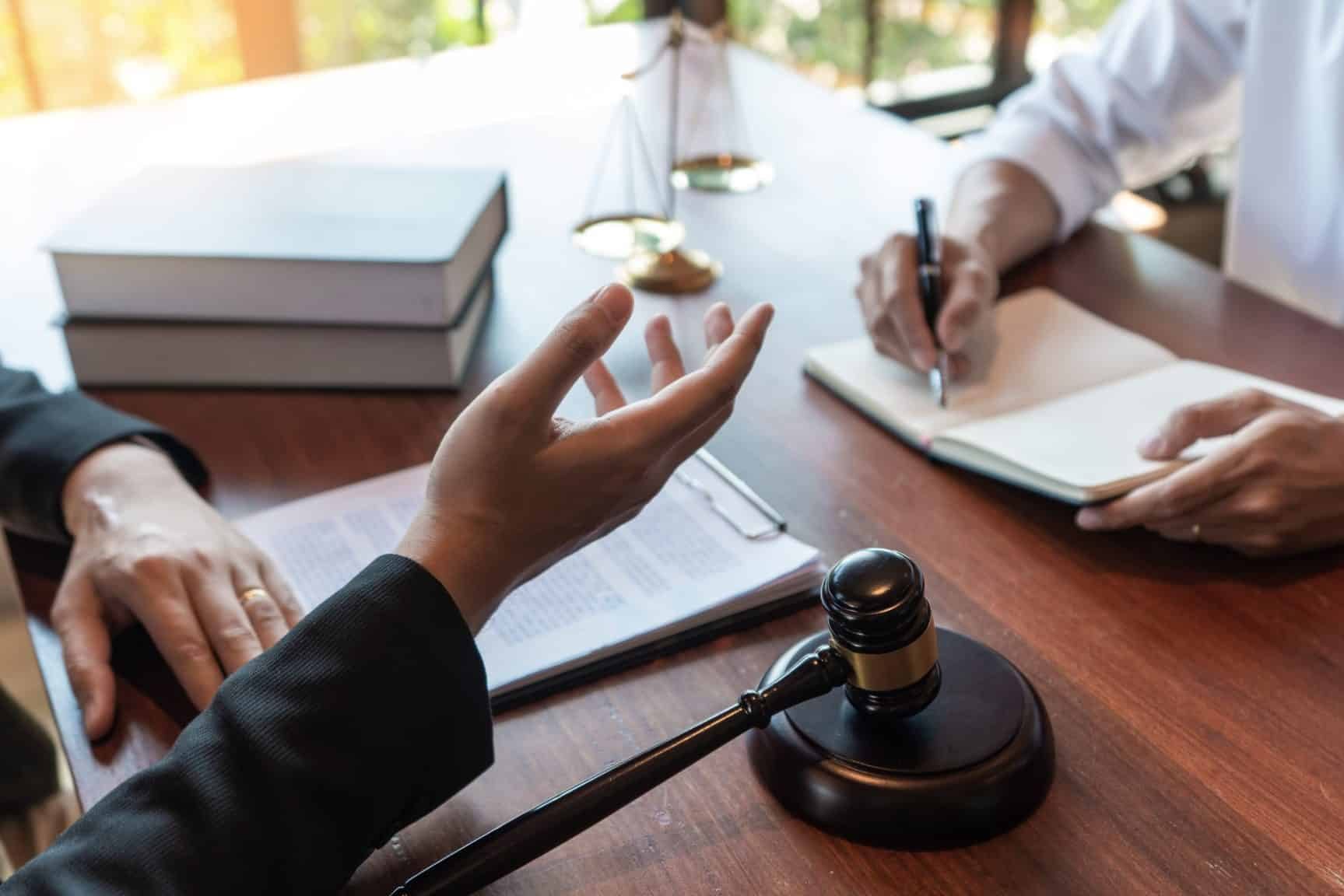 What Personal Injury Lawyers Actually Do After You Hire Them Two people sit at a desk with legal documents, a gavel, and scales of justice; one person gestures while the other takes notes.