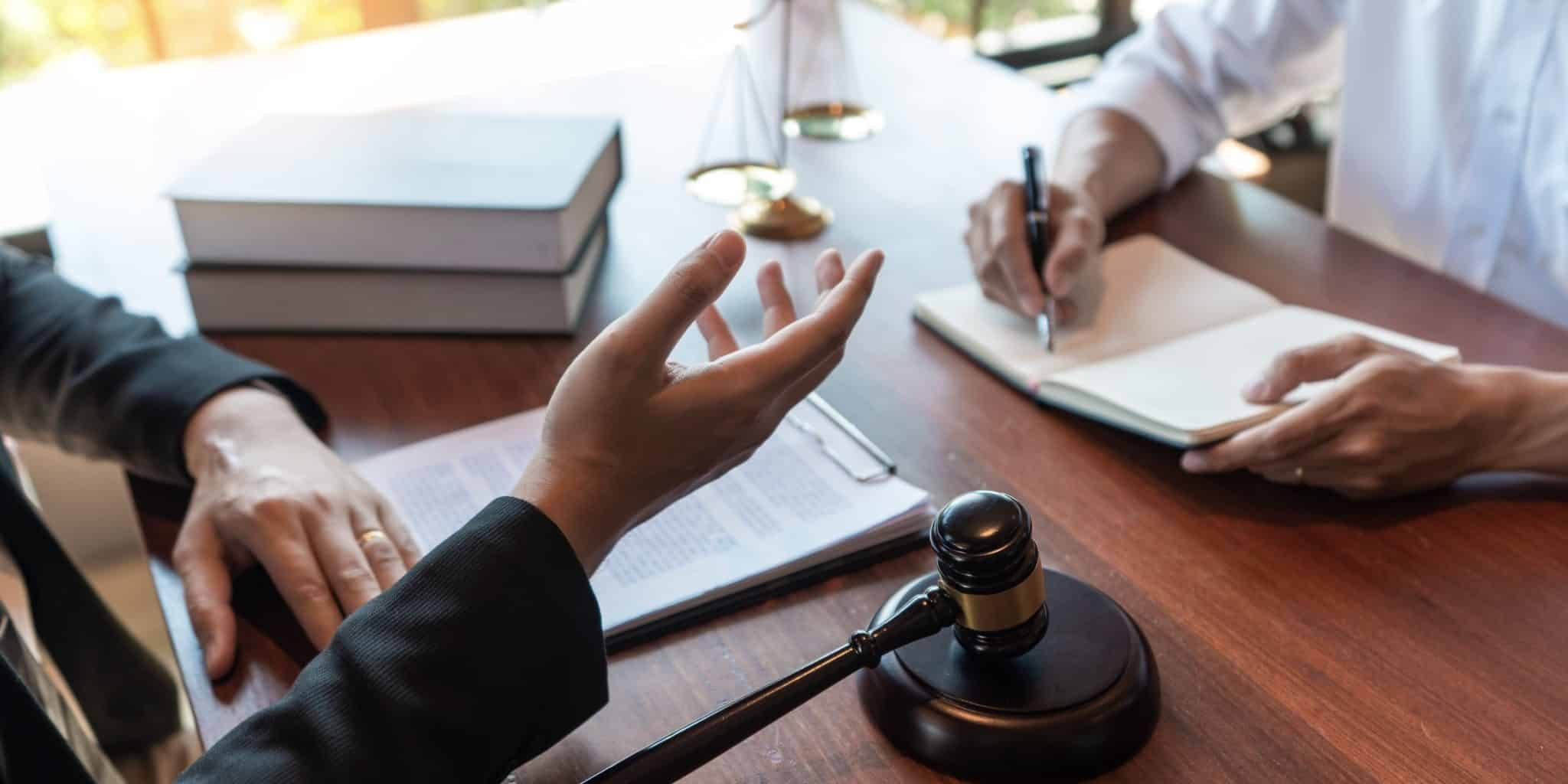 What Personal Injury Lawyers Actually Do After You Hire Them Two people sit at a desk with legal documents, a gavel, and scales of justice; one person gestures while the other takes notes.