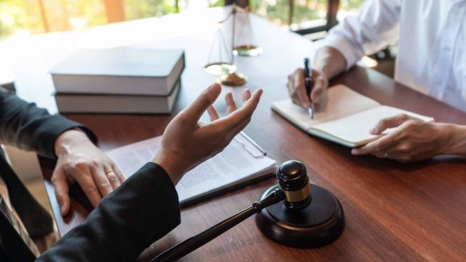 What Personal Injury Lawyers Actually Do After You Hire Them Two people sit at a desk with legal documents, a gavel, and scales of justice; one person gestures while the other takes notes.