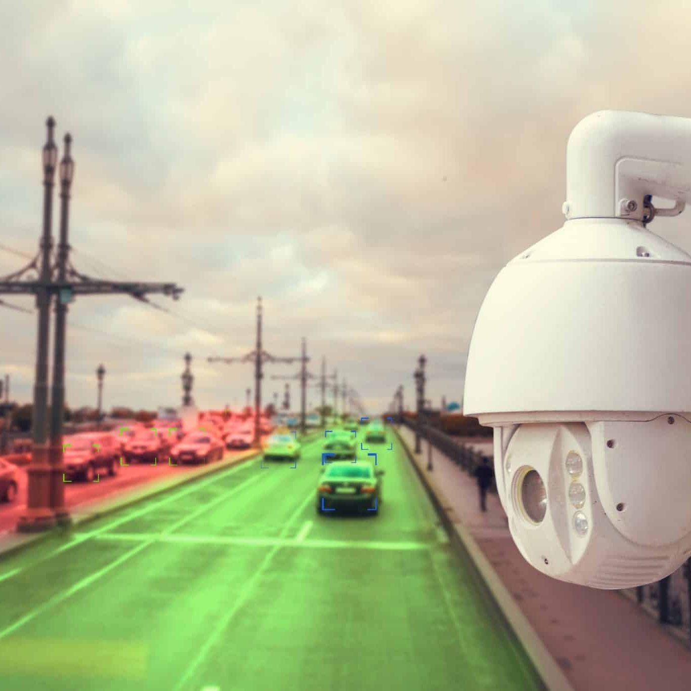 Surveillance camera overlooking a busy city bridge with cars; some vehicles highlighted in green and red, suggesting automated traffic monitoring.