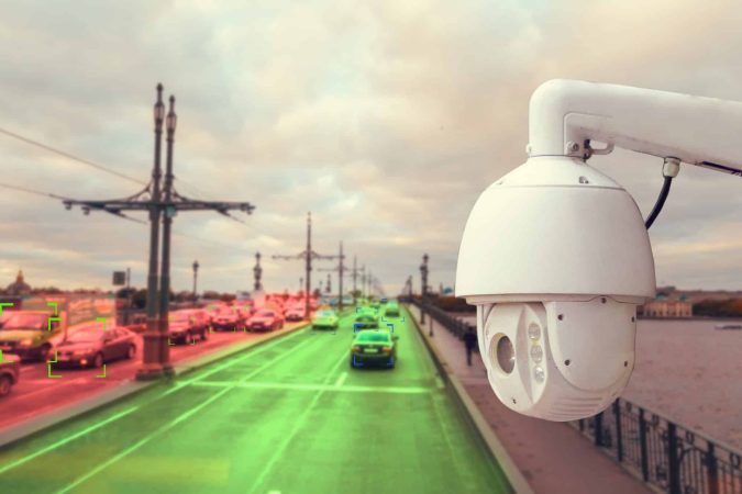 Surveillance camera overlooking a busy city bridge with cars; some vehicles highlighted in green and red, suggesting automated traffic monitoring.