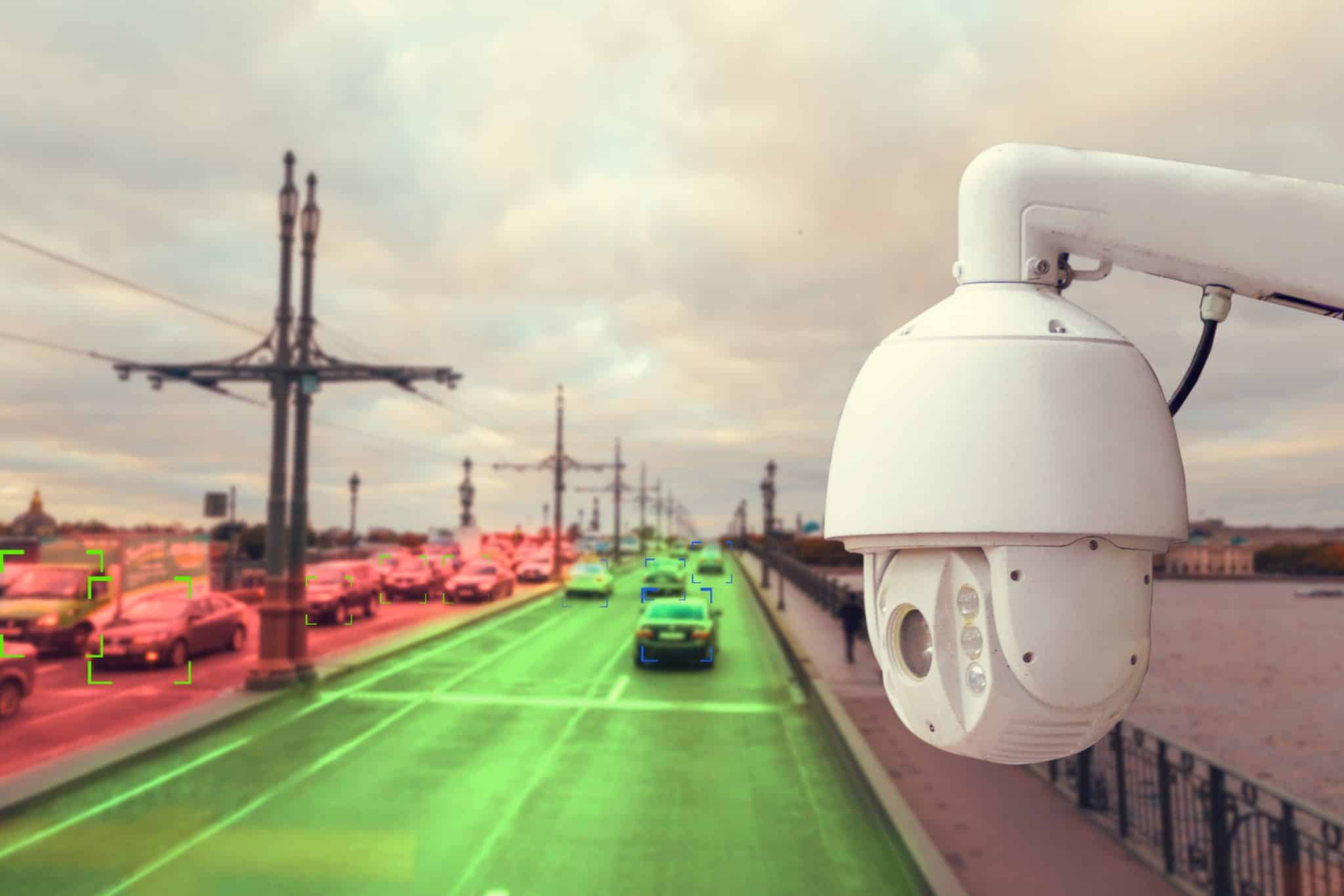 Surveillance camera overlooking a busy city bridge with cars; some vehicles highlighted in green and red, suggesting automated traffic monitoring.